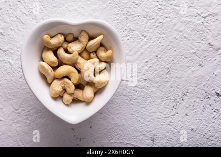 Top view, extreme close-up, against a textured white concrete background, a heart shaped bowl with cashew nuts. Copy space. Stock Photo
