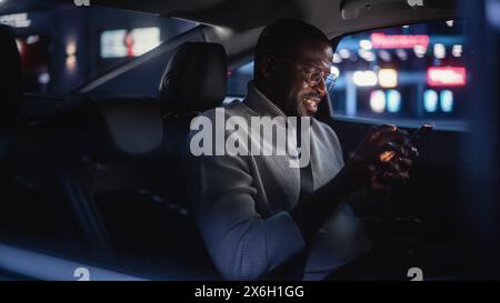 Happy Black Man in Glasses is Commuting Home in a Backseat of Taxi at Night. Handsome Male Using Smartphone and Smiling while in a Car in Urban City Street with Working Neon Signs. Stock Photo