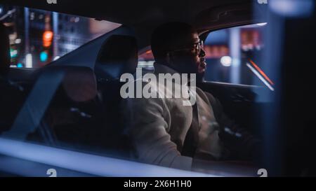 Stylish Black Man in Glasses is Commuting Home in a Backseat of a Taxi at Night. Handsome Male Passenger Looking Out of Window while in a Car in Urban City Street with Working Neon Signs. Stock Photo