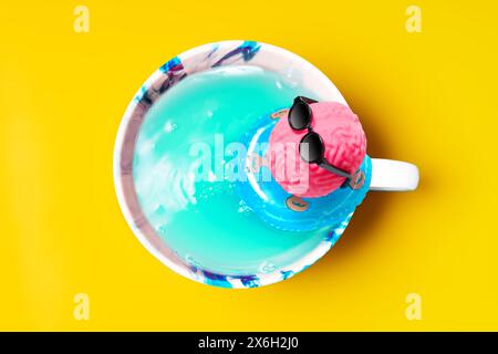 Impromptu pool in a mug, where a pink brain, adorned with sunglasses, lounges on an inflatable ring isolated on yellow background. Leisure, innovation Stock Photo
