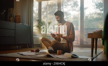 Man Using Smartphone at Home, Does Remote Work. Handsome Smiling Male Sitting on the Floor Works with Papers, Documents, Brainstorms Creative Project with Online Research. Sunny Living Room Stock Photo