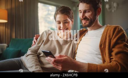 Couple Use Smartphone Device, while Sitting on a Couch in the Cozy Apartment. Boyfriend and Girlfriend Shopping on Internet, Watching Funny Videos, Use Social Media, Streaming Service. Stock Photo