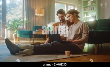 Couple Use Laptop Computer, while Sitting on the Living Floor room of their Apartment. Boyfriend and Girlfriend Talk, Shop on Internet, Choose Product to Order Online, Doing Remote Work Together Stock Photo