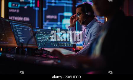 Professional IT Technical Support Specialist and Software Developer Working on Computer in Monitoring Control Room with Digital Screens. Employee Uses Headphones with Mic, Talking on a Call. Stock Photo