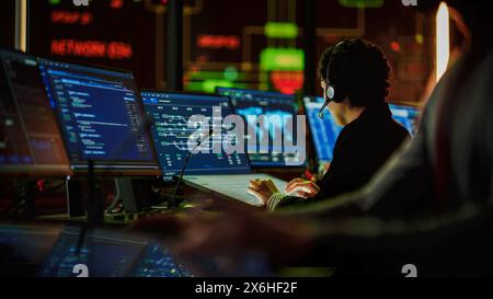 Multiethnic Female IT Technical Support Specialist Talking with a Client in Headphones and Working on Desktop Computer in Monitoring Control Room with Big Digital Screens with Server Blockchain Data. Stock Photo