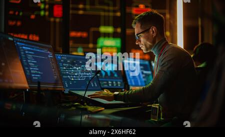 Professional IT Technical Support Specialist Making an Intercom Announcement on a Microphone and Working on Desktop Computer in Monitoring Control Room with Big Digital Screens with Server Data. Stock Photo