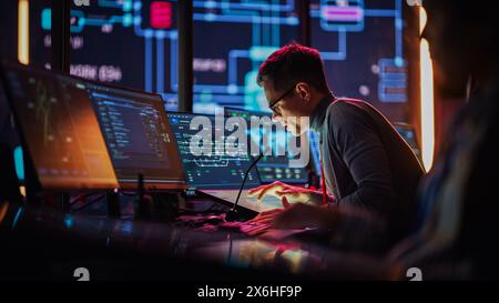 Professional IT Technical Support Specialist Making an Intercom Announcement on a Microphone and Working on Desktop Computer in Monitoring Control Room with Big Digital Screens with Server Data. Stock Photo