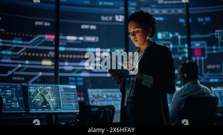 Young Multiethnic Female Government Employee Uses Tablet Computer in System Control Monitoring Center. In the Background Her Coworkers at Their Workspaces with Many Displays Showing Technical Data. Stock Photo