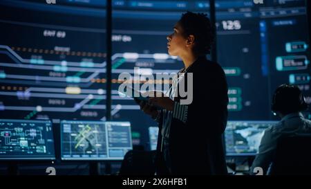 Young Multiethnic Female Government Employee Uses Tablet Computer in System Control Monitoring Center. In the Background Her Coworkers at Their Workspaces with Many Displays Showing Technical Data. Stock Photo