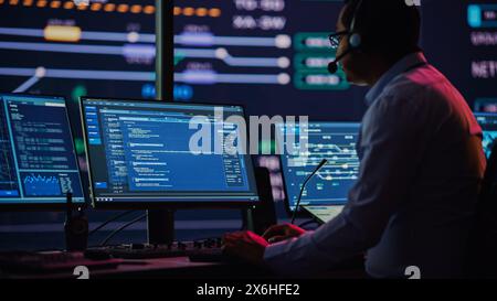 Professional IT Technical Support Specialist and Software Developer Working on Computer in Monitoring Control Room with Digital Screens. Employee Uses Headphones with Mic and Talking on a Call. Stock Photo