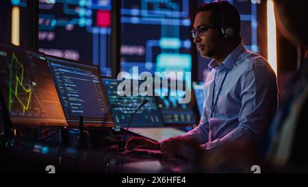 Professional IT Technical Support Specialist and Software Developer Working on Computer in Monitoring Control Room with Digital Screens. Employee Uses Headphones with Mic, Talking on a Call. Stock Photo