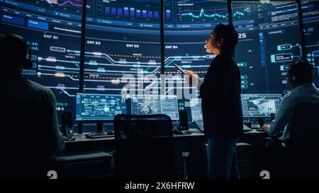 Young Multiethnic Female Government Employee Uses Tablet Computer in System Control Monitoring Center. In the Background Her Coworkers at Their Workspaces with Many Displays Showing Technical Data. Stock Photo