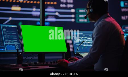 Professional IT Technical Support Specialist and Software Developer Working on Computer with Green Screen Mock Up Display in Monitoring Control Room with Digital Screens. Employee Uses on Headphones. Stock Photo