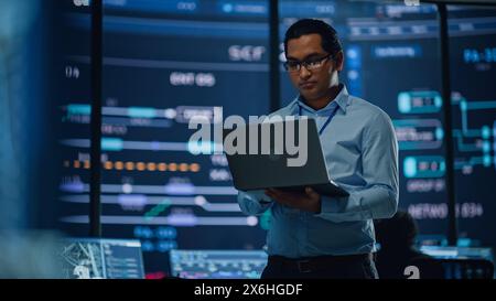 Young Multiethnic Male Government Employee Uses Laptop Computer in System Control Monitoring Center. In the Background His Coworkers at Their Workspaces with Many Displays Showing Technical Data. Stock Photo