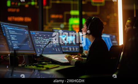 Multiethnic Female IT Technical Support Specialist Talking with a Client in Headphones and Working on Desktop Computer in Monitoring Control Room with Big Digital Screens with Server Blockchain Data. Stock Photo