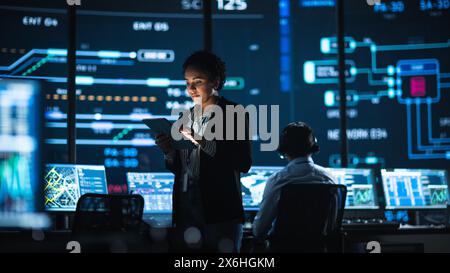 Young Multiethnic Female Government Employee Uses Tablet Computer in System Control Monitoring Center. In the Background Her Coworkers at Their Workspaces with Many Displays Showing Technical Data. Stock Photo