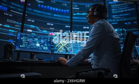 Professional IT Technical Support Specialist and Software Developer Working on Computer in Monitoring Control Room with Digital Screens. Employee Uses Headphones with Mic and Talking on a Call. Stock Photo