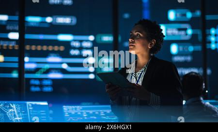 Young Multiethnic Female Government Employee Uses Tablet Computer in System Control Monitoring Center. In the Background Her Coworkers at Their Workspaces with Many Displays Showing Technical Data. Stock Photo