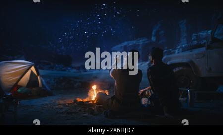 Happy Couple Tent Camping in the Canyon, Sitting by Campfire Watching Night Sky with Milky Way Full of Bright Stars. Two Travelers In Love On a Romantic Vacation Trip. Back View Shot Stock Photo