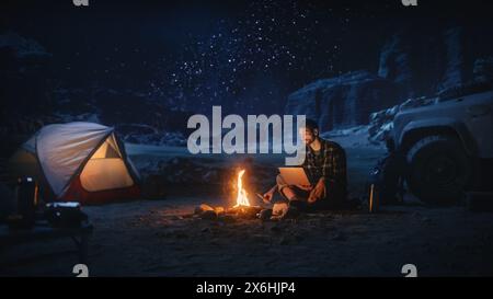 Male Traveler Sitting by Campfire Uses Laptop Computer while Tent Camping in the Canyon. Man doing Digital Remote Work, e-business, e-shopping, ecommerce through Internet while being on Vacation Trip Stock Photo