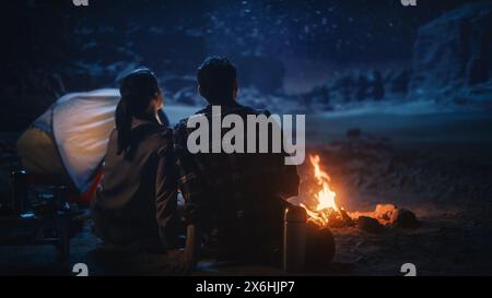 Happy Couple Tent Camping in the Canyon, Sitting by Campfire Watching Night Sky with Milky Way Full of Bright Stars. Two Travelers In Love On a Romantic Vacation Trip. Back View Shot Stock Photo