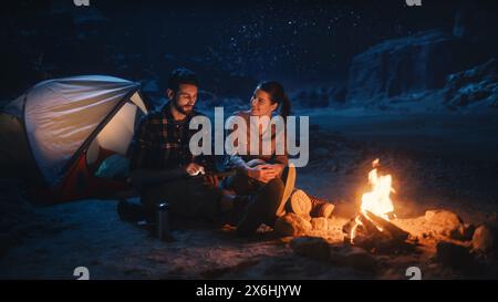 Happy Couple Nature Camping in the Canyon at Night, Use Digital Tablet Computer, Sitting by Campfire. Two people Have Fun, Smile, Post Traveling Photos on Social Media, Watch Funny Videos on Internet Stock Photo