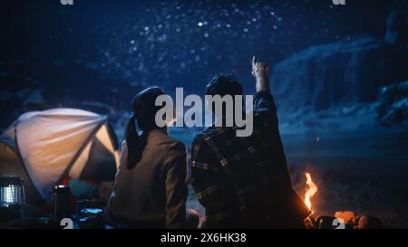 Happy Couple Tent Camping in the Canyon, Sitting by Campfire Watching Night Sky with Milky Way Full of Bright Stars. Two Travelers In Love On a Romantic Vacation Trip. Back View Shot Stock Photo