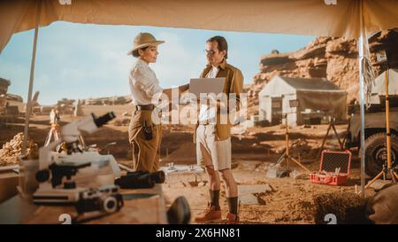 Archaeological Digging Site: Two Great Archeologists Stand in Tent Work on Excavation Site, Use Laptop Computer to Inspect Newly Discovered Ancient Civilization Architectural Site, Planning Work Stock Photo