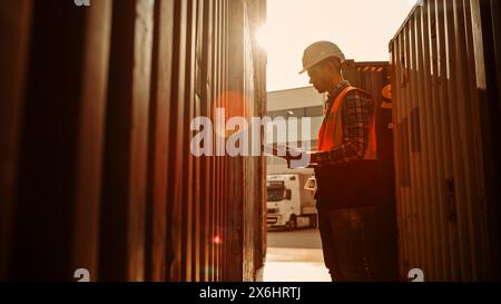 Handsome Caucasian Industrial Inspector or Supervisor Wearing White Hard Hat and High-Visibility Vest is Examining the Containers with His Tablet Computer in a Logistics Shipping Terminal Depot. Stock Photo