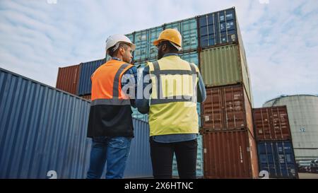 Handsome Industrial Engineer and African American Supervisor in Hard Hats and Safety Vests Standing with Their Backs to the Camera in Container Terminal. Colleagues Talk About Logistics Operations. Stock Photo