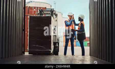 Forklift Driver Loading a Shipping Cargo Container with a Full Pallet with Boxes in Logistics Port Terminal. Latin Female Industrial Supervisor and Safety Inspector with Tablet Giving Him High Five. Stock Photo