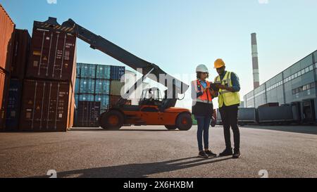 Multiethnic Female Industrial Engineer with Tablet and Black African American Male Supervisor in Hard Hats and Safety Vests Stand in Container Terminal. Colleagues Talk About Logistics Operations. Stock Photo