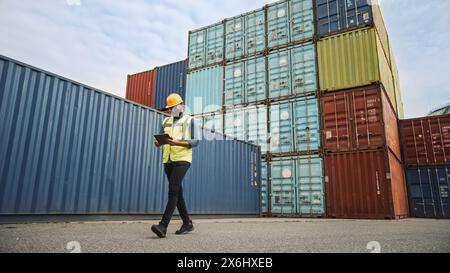 Black African American Male Industrial Supervisor in Hard Hat and Safety Vest Talking is Using Tablet Computer and Walking Next to a Container in Logistics Operations Terminal. Stock Photo