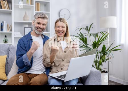 Joyful couple cheering while using a laptop, seated on sofa in cozy, modern living room. Expressing success and happiness. Stock Photo