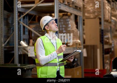 Young caucasian man worker in hard hat and working wear outfit doing stocktaking of products on shelves in warehouse using clipboard, taking notes. Ph Stock Photo