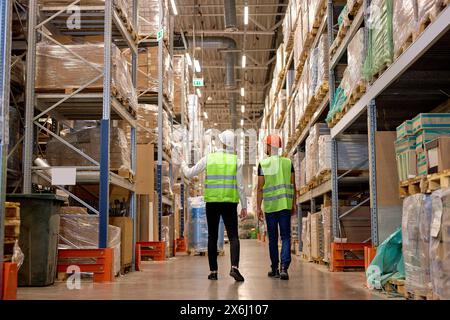Two warehouse workers in vest and helmet walking in distribution storage area discussing, in workplace area. Concept of good management system to supp Stock Photo