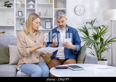 Middle-aged couple reviewing financial documents at home on the couch, looking concerned. Concept of financial planning, budgeting, and dealing with paperwork. Stock Photo