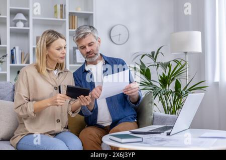 Mature couple sitting on a couch, reviewing documents and using a tablet at home. They seem focused and engaged in a financial discussion. Stock Photo