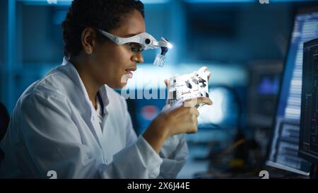 Modern Electronics Research, Development Facility: Black Female Engineer Wearing High-Tech Glasses Inspects Printed Circuit Board Motherboard. Scientist Designs Silicon Microchips, Semiconductors Stock Photo