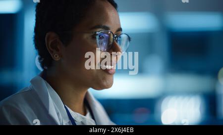 Beautiful Black Latin Woman Wearing Glasses Smiling Charmingly. Young Intelligent Female Scientist Working in Laboratory. Background Bokeh Blue with High-Tech Technological Lights Stock Photo