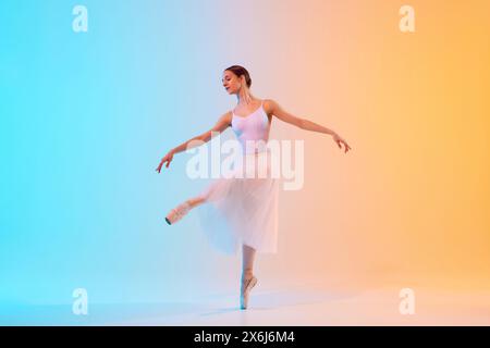 Young ballerina in white tutu an pointe practicing dance moves in neon light against blue-orange gradient background. Stock Photo