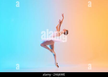 Young ballerina in white tutu dancing and leaning backwards in motion in neon light against blue-orange gradient background. Stock Photo