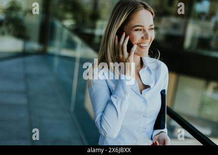 A young businesswoman smiles confidently while talking on her phone inside a sleek, modern office building, with cityscape reflections in the glass. Stock Photo