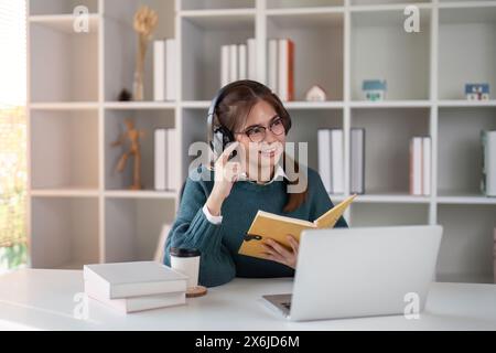 Young woman wearing headphones while studying with a book and laptop at a home office desk Stock Photo