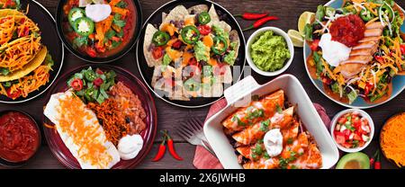 Mexican food table scene. Top down view on a dark wood banner background. Tacos, burrito plate, nachos, enchiladas, tortilla soup and salad. Stock Photo