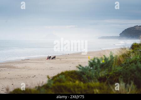 Man and woman sitting on the beach watching the sunset, rocky landscape in the background. Taken at Waihi Beach in New Zealand Stock Photo