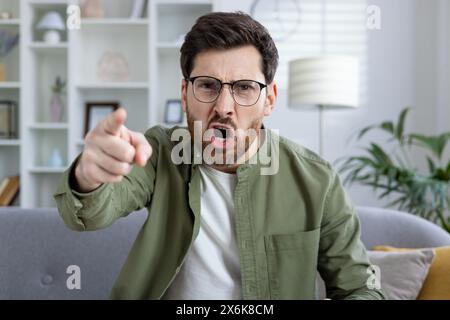 Angry man wearing glasses and a green shirt pointing and yelling while sitting on a couch in a modern living room with shelves ,in the background. Stock Photo