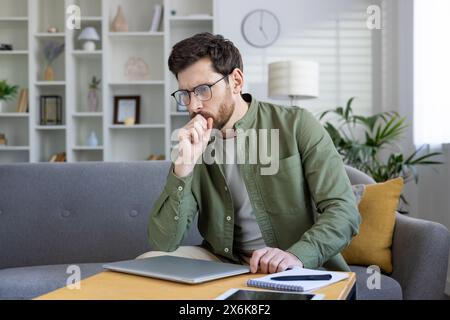 Thoughtful young man working on his laptop at home. He is thinking and planning while sitting on the sofa. Stock Photo