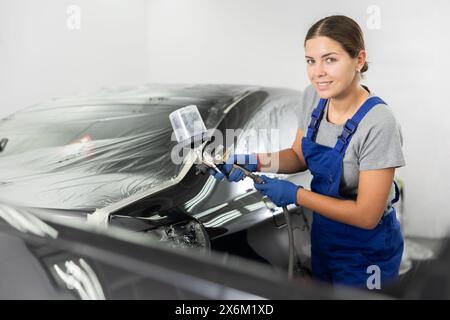 Car painting and automobile repair service. Woman in blue overalls paints car with airbrush pulverizer in paint chamber Stock Photo