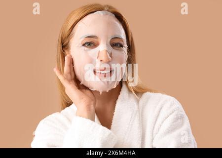 Mature woman in bathrobe with facial sheet mask on beige background, closeup Stock Photo